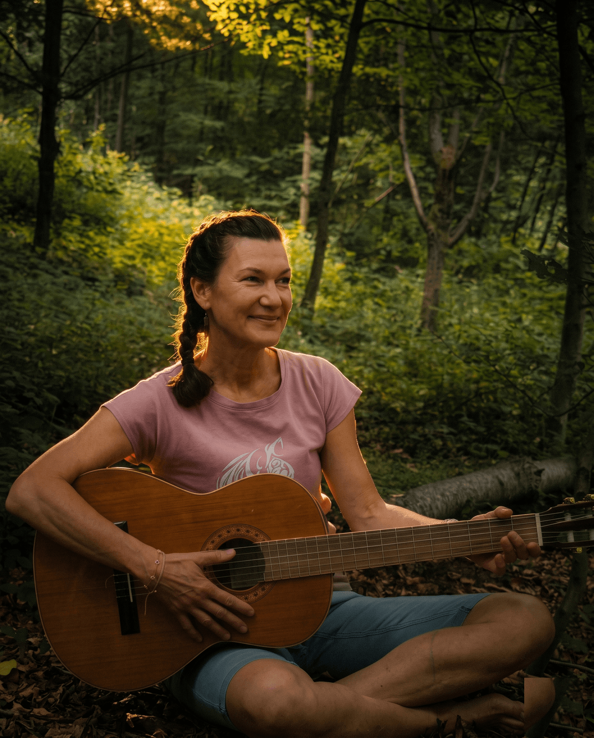 Heike sitzt im Schneidersitz auf dem Waldboden, spielt lächelnd eine Akustikgitarre, goldenes Abendlicht zwischen den Bäumen
