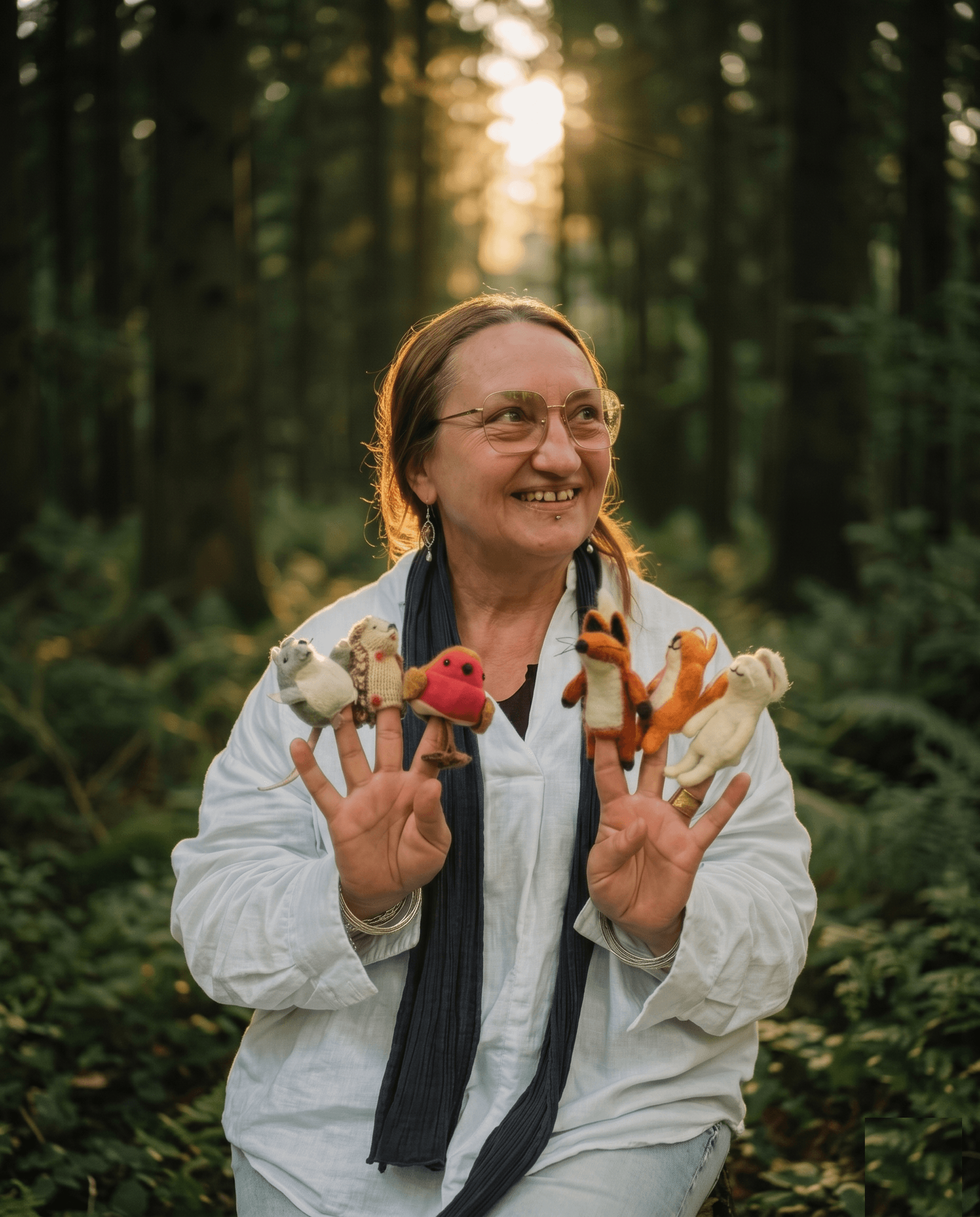 Mina lächelnd in weisser Leinenbluse, hält beide Hände mit kleinen Tier-Fingerpuppen hoch, tief stehende Sonne im Wald