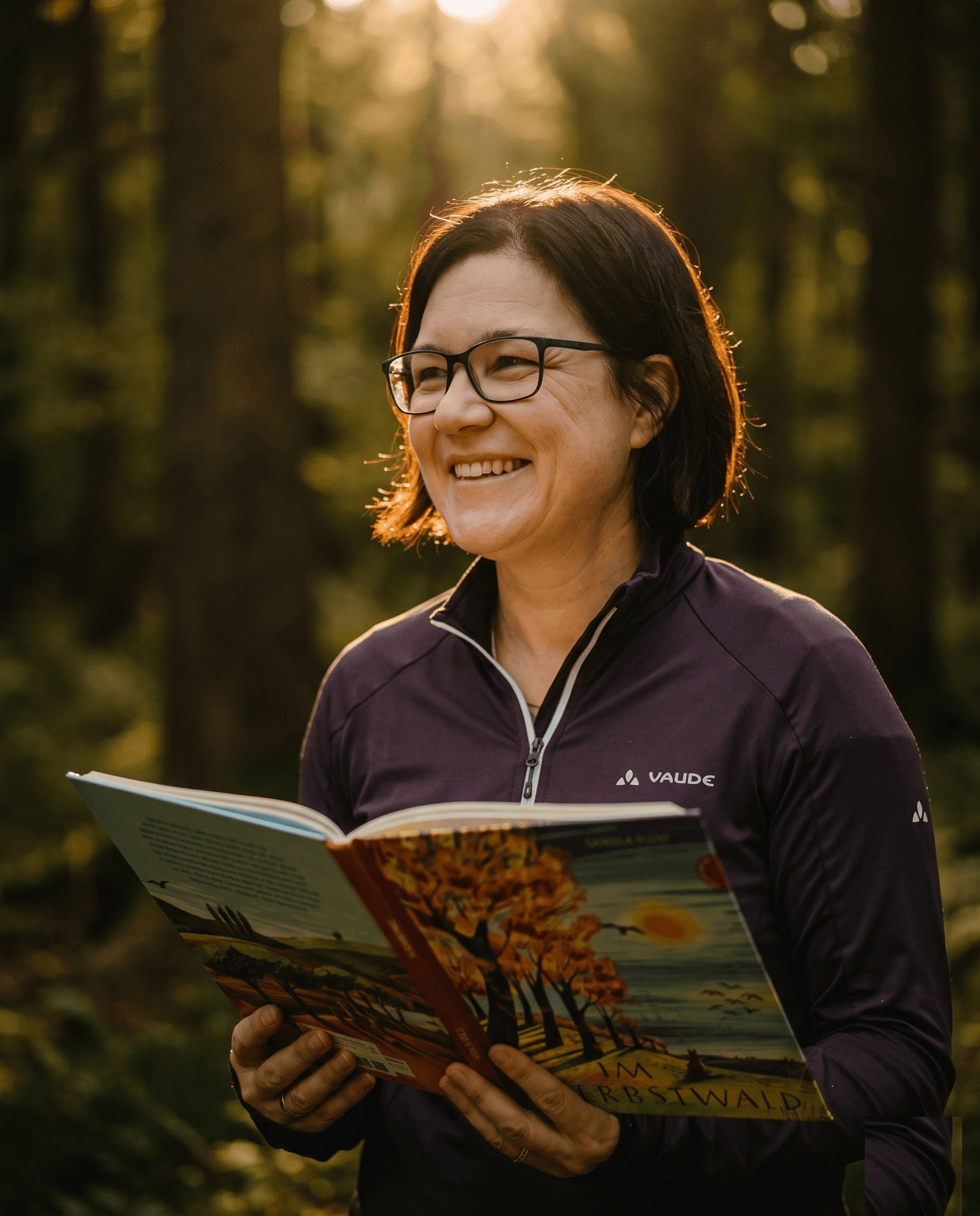 Nicole in Fleecejacke und Brille, lächelnd mit einem aufgeschlagenen Bilderbuch, dahinter sonnendurchfluteter Wald